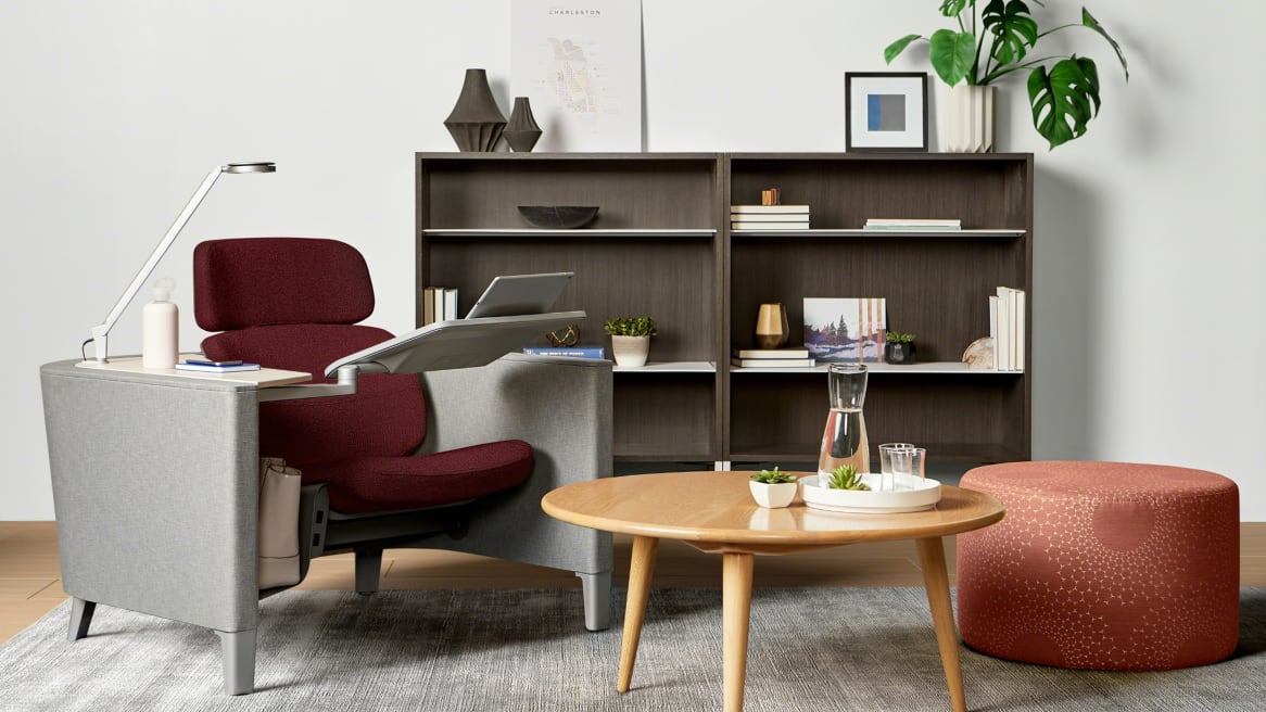 Private room with a red Brody seat with a Dash light and personal table, a laptop on it. A wooden coffee table with a water vase on it. A wooden storage behind with a plant above it.